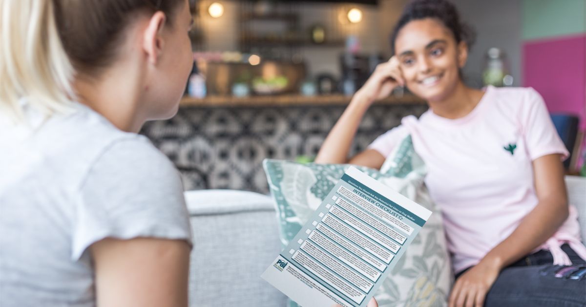 A pet owner in a white t-shirt sits on her couch holding a pet sitter interview checklist. The pet sitter is sitting near her on the other side of the couch.